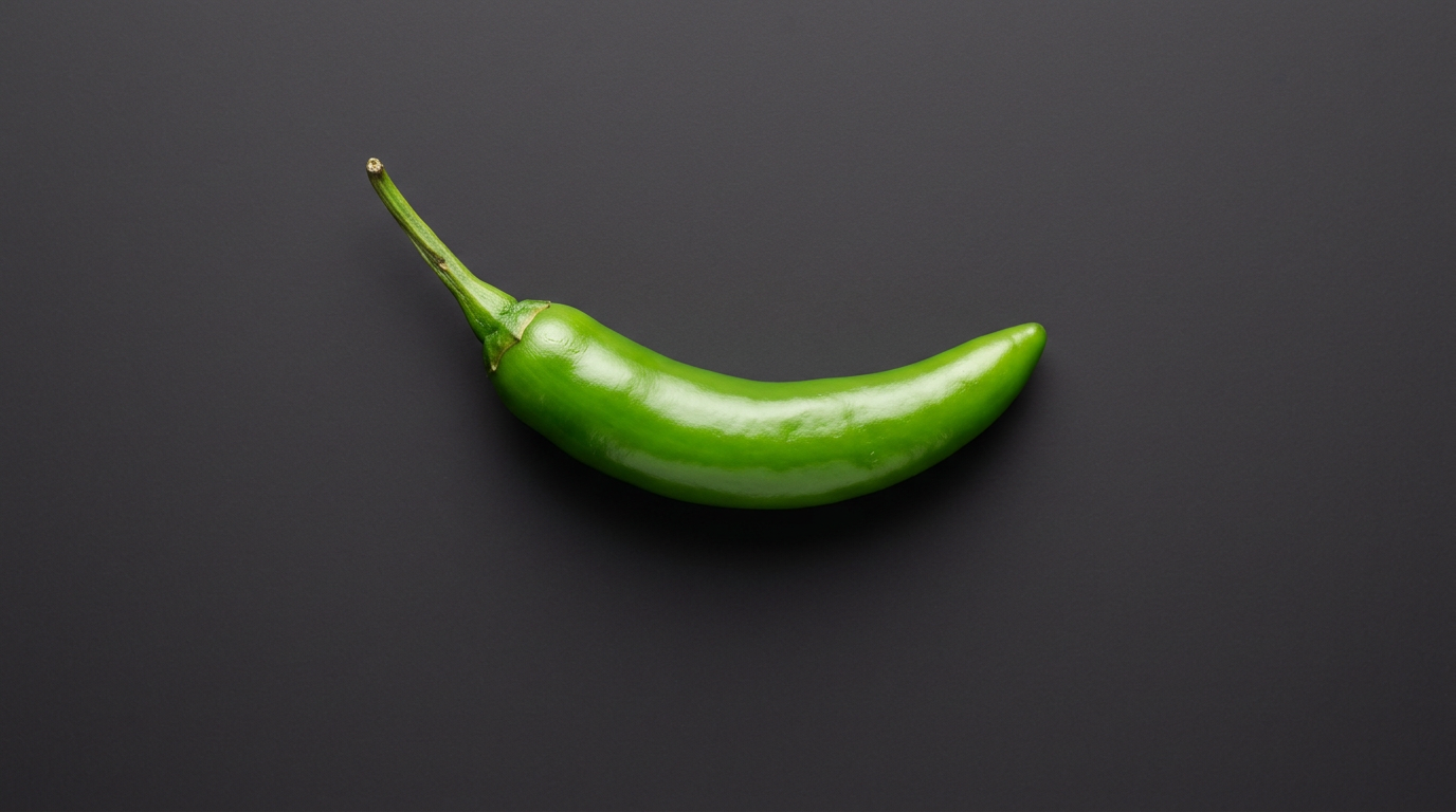 A single fresh green Serrano pepper, smaller and thinner than a Jalapeño, photographed against a dark charcoal background.